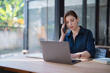 Fototapeta premium Businesswoman multitasking, engaging in a phone call while simultaneously working on a laptop at a desk, demonstrating efficient remote work and communication
