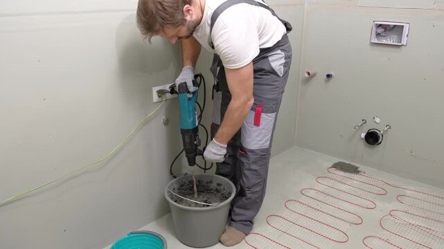 Construction worker preparing tile surface, mixing mortar substrate for flooring, underfloor heating mat. Workman combines adhesive in renovation setting. Operating power tool during bathroom upgrade