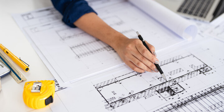 Close up hand of Architect or engineer hand holding pen working on blueprint construction floor plans on desk with engineering tools.