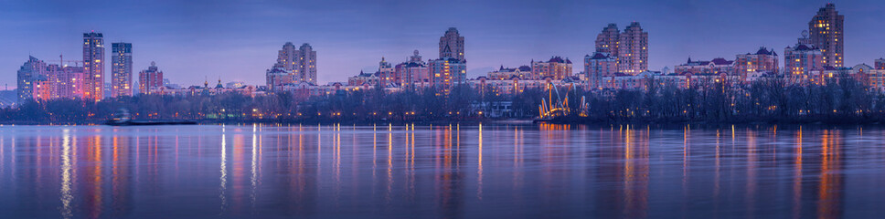 Night over Obolon embankment in Kyiv with modern residential skyline with lights reflected in calm river water, dramatic sky creating a peaceful urban evening atmosphere.