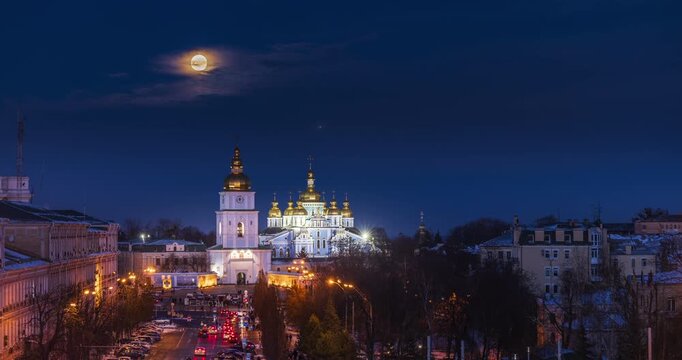Winter evening at Sofia Square in Kyiv with the rising moon above St. Michael&rsquo;s Golden-Domed Monastery, glowing domes, snowy streets, city lights, calm blue sky, historic urban atmosphere.