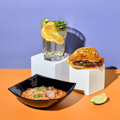 Mexican cuisine dishes, set meal of tacos, birria ramen soup, corn nachos chips and lemonade on colored background