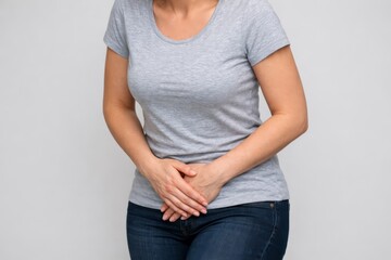 Woman in casual clothing holding lower abdomen with hands, depicting stomach pain, cramps, digestive discomfort, or pelvic ache on neutral background