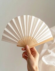 Delicate Hand Holds Open White Paper Folding Fan with Light Wood Handle Against Soft White Fabric Background and Gentle Natural Light