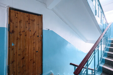 Wooden door next to light blue-painted staircase with metal railing; concrete steps, white upper walls, utilitarian residential hallway design