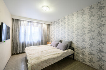 Fototapeta premium Neat bedroom with double bed, floral wallpaper, wall-mounted TV, sheer curtains, wooden bedside table, soft lighting, light-colored flooring, cozy pillows