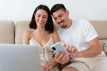 Happy couple engaging with digital media on a smartphone and laptop while relaxing on a sofa, sharing a moment and enjoying online activities together at home