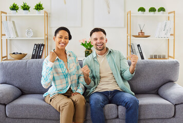 Happy biracial couple African woman and Caucasian man on sofa at home watching soccer match sports live stream, celebrating goal, cheering with fists raised, sharing moment of joy and triumph together
