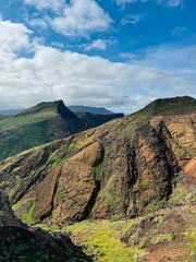Madeira Ponta de Sao Lourenco A rugged mountain landscape with reddish rocky terrain, green slopes, and striking peaks stretches under a blue sky with white clouds.