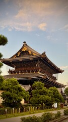 A monumental wooden temple gate rises majestically into the sky. Surrounded by green trees, the traditional Japanese architecture reveals complex details.