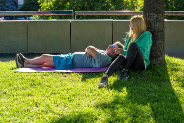 Middle aged man lying on a mat while a woman uses her phone by a tree