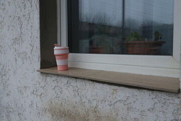 Coffee cup sits on a windowsill next to a house with plants in pots outside on a cloudy day
