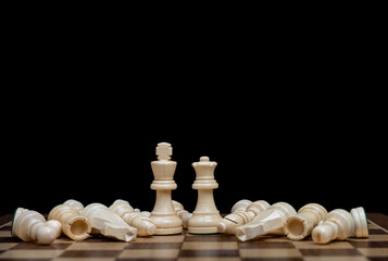 White King and Queen chess pieces stand tall on a wooden chessboard surrounded by fallen opponents against a stark black background