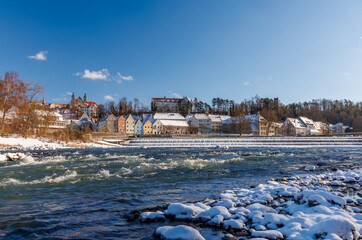 Scenic winter panorama of a historic European town by a river. Beautiful landscape featuring a large weir, snow-covered riverbanks, and a skyline of traditional colorful houses under a bright blue sky