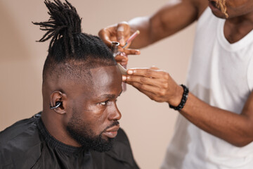 Close-up studio image of a professional barber using an airbrush tool and card to define the hairline of a Black adult man with short dreadlocks and a beard wearing a black barber cape.
