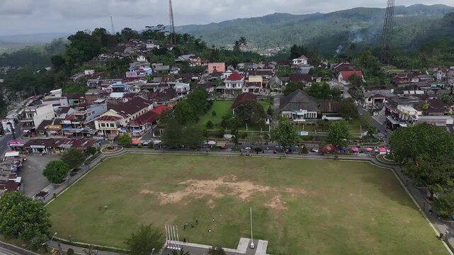Aerial perspective of Magelang, Indonesia, highlighting a vast central field within a residential town, framed by green hills under an overcast sky