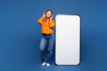Full body young happy woman in yellow sweater casual clothes big huge blank screen mobile cell phone smartphone with area listen music in headphones isolated on plain blue background studio portrait