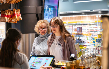 Two women smiling while ordering drinks and interacting with a cashier at a retail counter inside a brightly lit modern airport store