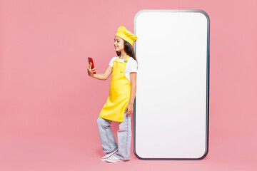 Full body small kid child girl housewife chef cook baker mother helper wear yellow apron cap big huge blank screen area use mobile cell phone isolated on plain pink background. Cooking food concept.
