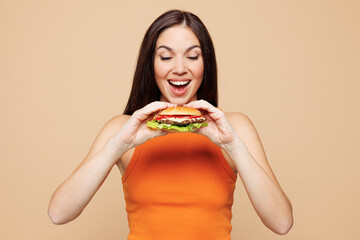 Young smiling happy cool woman wear orange casual clothes hold eat look at tasty burger isolated on plain pastel light beige background. Proper nutrition healthy fast food unhealthy choice concept
