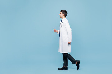 Full body side view young happy male doctor man wear white medical gown suit work in hospital clinic office walk go run isolated on plain blue background studio portrait. Healthcare medicine concept.