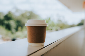 A coffee cup sits on a balcony railing. The cup is brown with a white lid. The blurred background reveals natural light and the green of the trees, creating a simple and relaxing atmosphere, perfect f