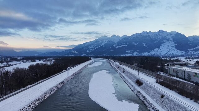Drone flying over the snow-covered Rhine River between Liechtenstein and Switzerland, showing a sandbank in the middle of the water partially covered in white snow.