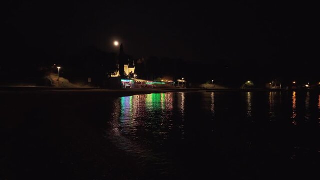 The full moon and colourful night lights of a beach bar reflecting on the calm sea surface in Njivice, Croatia. Njivice is a coastal village located on Krk Island in Primorje-Gorski Kotar County.
