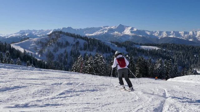 Caucasian skier carving alpine slope panoramic mountain vista. View on pine forest below and distant high summits crisp sunlight glinting on fresh powder snow. Skier smooth turns on ski slope
