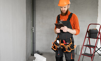 male construction worker or builder in hard hat, tool belt and overall with tablet pc computer in the room. Concept of profession, construction and building