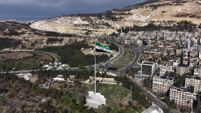 Aerial View of Syrian National Flag Waving on High Flagpole with Mount Qasioun and Damascus Cityscape in Background