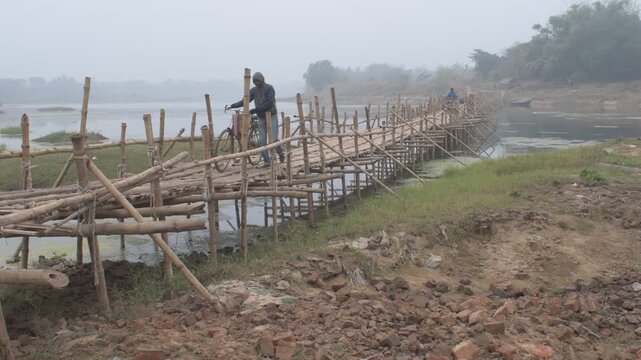 a traditional handmade bamboo bridge spanning calm, misty 'kangshabati" river in a rural landscape, showing local unrecognized people crossing on foot and motorcycle