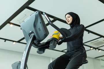Muslim women in hijab using stationary exercise bike at gym, holding yellow water bottle. Promotes cardio fitness, hydration, active lifestyle. Great for health, gym ads and inclusive wellness.