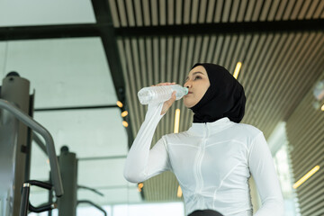 Muslim woman in white sportswear drinking water from bottle at gym. Highlights hydration, post workout recovery, wellness lifestyle. Ideal for fitness ads, health brands and hijab sports themes.