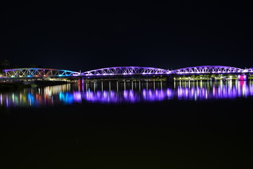The historic Truong Tien Bridge radiates purple light across the river at night, hovering above a passing traditional dragon boat