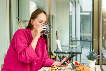 A young Caucasian woman with long brown hair drinks coffee while using a tablet. She wears a pink shirt and is seated at a table with breakfast items.