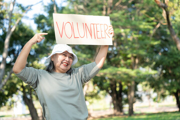 A senior woman smiles while holding a VOLUNTEER sign in a sunny park. The image highlights active aging, community service, and the spirit of giving back through outdoor social engagement