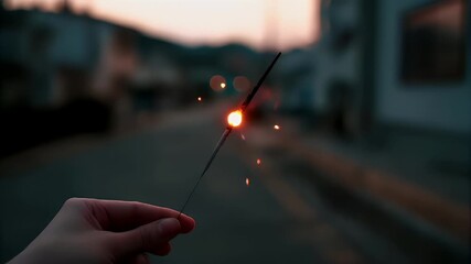 A hand holding a sparkler against a backdrop of a sunset over a city street. The sparkler is the central focus, with its bright, fiery glow illuminating the scene.