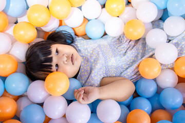 Adorable Little Asian Girl Relaxing and Lying in Colorful Plastic Ball Pit,A cute young Asian girl lying down and covered with many blue, orange, and white plastic balls in a playground ball pit,