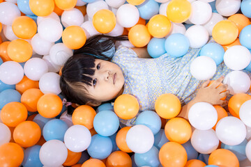 Adorable Little Asian Girl Relaxing and Lying in Colorful Plastic Ball Pit,A cute young Asian girl lying down and covered with many blue, orange, and white plastic balls in a playground ball pit,