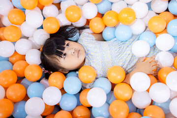 Adorable Little Asian Girl Relaxing and Lying in Colorful Plastic Ball Pit,A cute young Asian girl lying down and covered with many blue, orange, and white plastic balls in a playground ball pit,