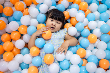 Adorable Little Asian Girl Relaxing and Lying in Colorful Plastic Ball Pit,A cute young Asian girl lying down and covered with many blue, orange, and white plastic balls in a playground ball pit,