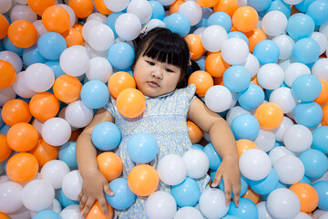 Adorable Little Asian Girl Relaxing and Lying in Colorful Plastic Ball Pit,A cute young Asian girl lying down and covered with many blue, orange, and white plastic balls in a playground ball pit,