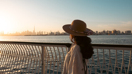 young woman wearing a hat and flowy dress exploring Dubai with Dubai skyline background, she is walking at the Creek harbor waterfront promenade at sunset