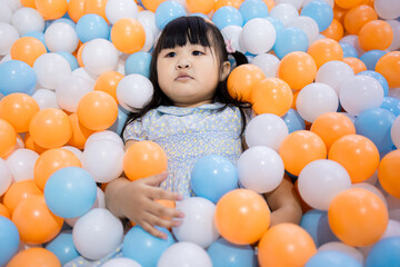 Adorable Little Asian Girl Relaxing and Lying in Colorful Plastic Ball Pit,A cute young Asian girl lying down and covered with many blue, orange, and white plastic balls in a playground ball pit,
