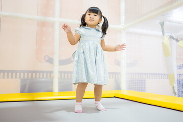 Cute Little Asian Girl Jumping and Playing on Trampoline in Indoor Playground,A cheerful young Asian girl in a blue dress enjoying her time jumping on a trampoline at an indoor soft play center.