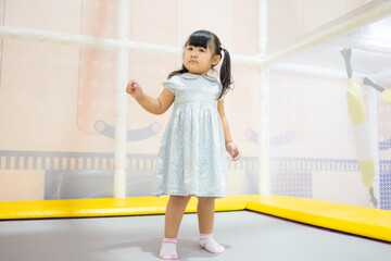 Cute Little Asian Girl Jumping and Playing on Trampoline in Indoor Playground,A cheerful young Asian girl in a blue dress enjoying her time jumping on a trampoline at an indoor soft play center.