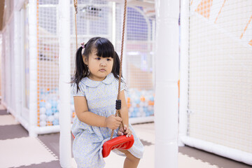 Little Asian girl playing on a swing in an indoor playground,A cute young Asian girl with pigtails sitting on a red swing, looking focused while playing in a colorful indoor playroom or kids club.