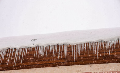 Long icicles hanging from a snow-covered roof during active snowfall, illustrating winter weather hazards, freezing temperatures and seasonal risks on residential buildings.