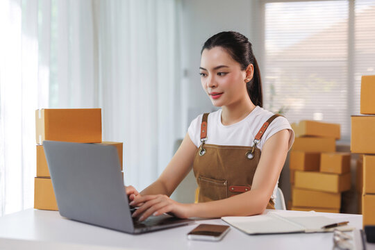 Young female entrepreneur wearing apron, using laptop for online store orders, with shipping boxes in a home office - Powered by Adobe